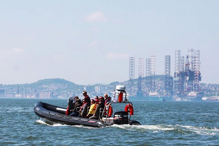 People on a black inflatable boat on water, with an oil rig and city in the background.