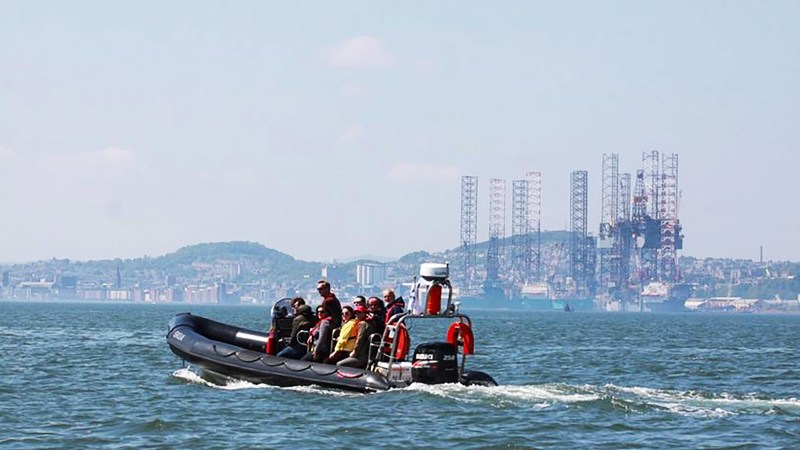 People on a black inflatable boat on water, with an oil rig and city in the background.