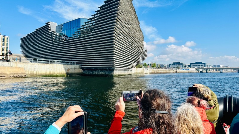 People on a boat photographing a modern, angular building by the water.