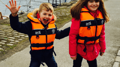 Two children wearing orange life jackets pose excitedly by a dock with boats.