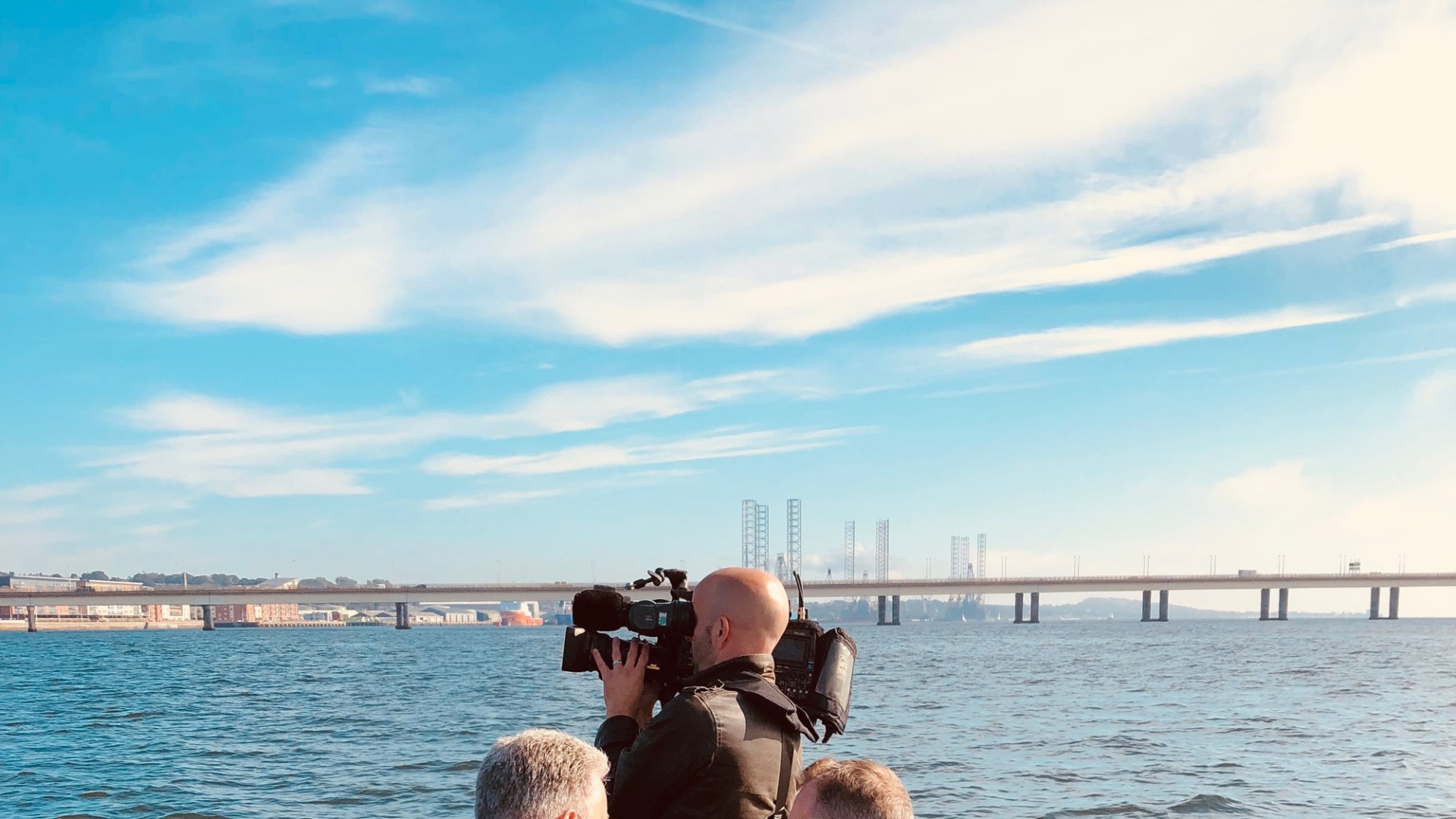People on a boat filming a wide bridge over a body of water on a clear day.