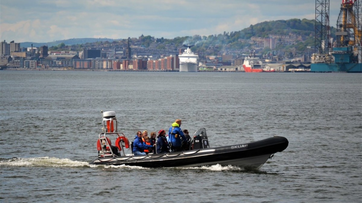 A speedboat with passengers on a river, city and hills in the background.