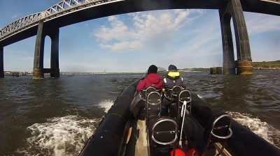 Two people on a boat approaching a large bridge over a river under a blue sky.