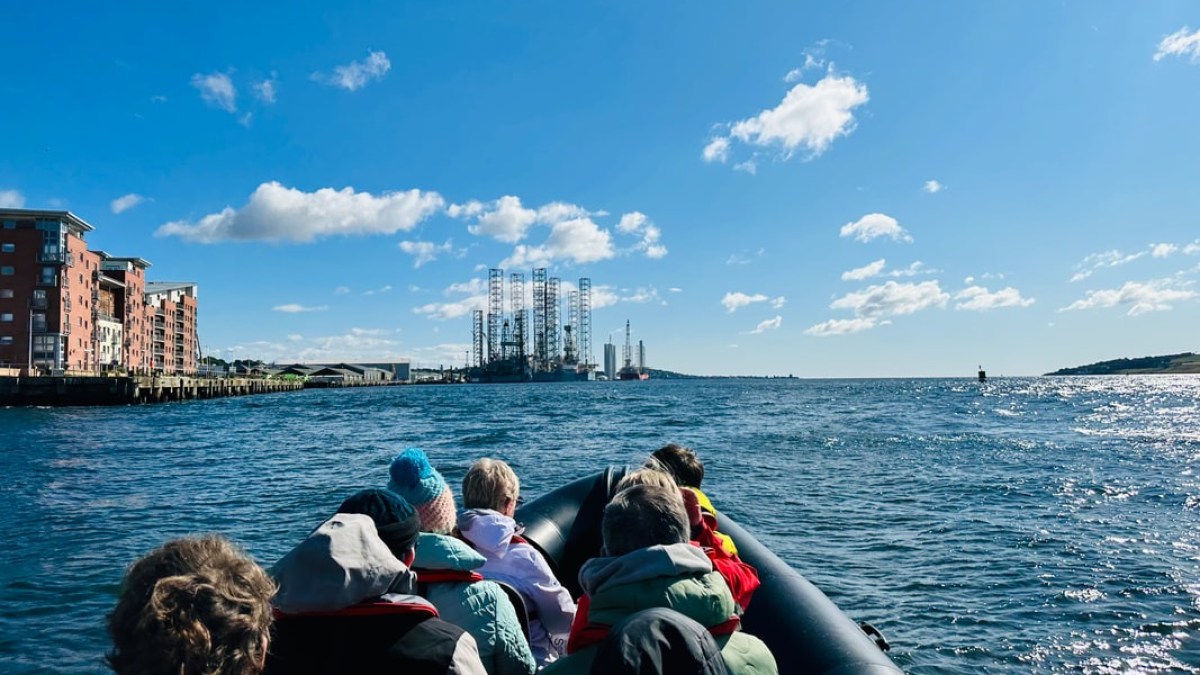 People on a boat heading towards an oil rig in the ocean, with buildings on the left and a blue sky.
