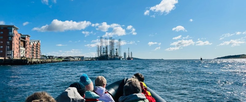 People on a boat heading towards an oil rig in the ocean, with buildings on the left and a blue sky.