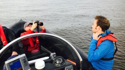 Two people in a boat, one taking a photo, the other wearing a blue jacket, with water and bridge in the background.