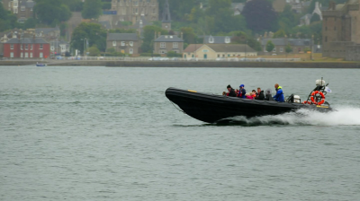 A speedboat with passengers moving quickly on a body of water near a shoreline with buildings.