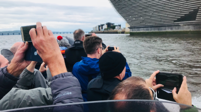 People on a boat taking photos of a modern, slanted building by the water.