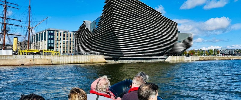 Group on boat viewing modern waterfront buildings under blue sky.