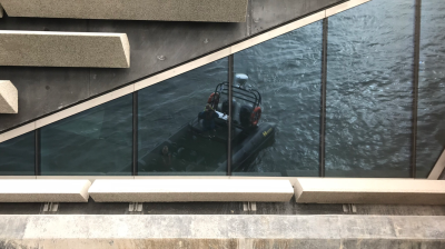 Glass facade reflects a boat on water, under a structure with diagonal concrete beams.
