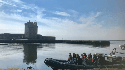 Group on a boat by a castle, calm waters, cobbled shore, blue sky.